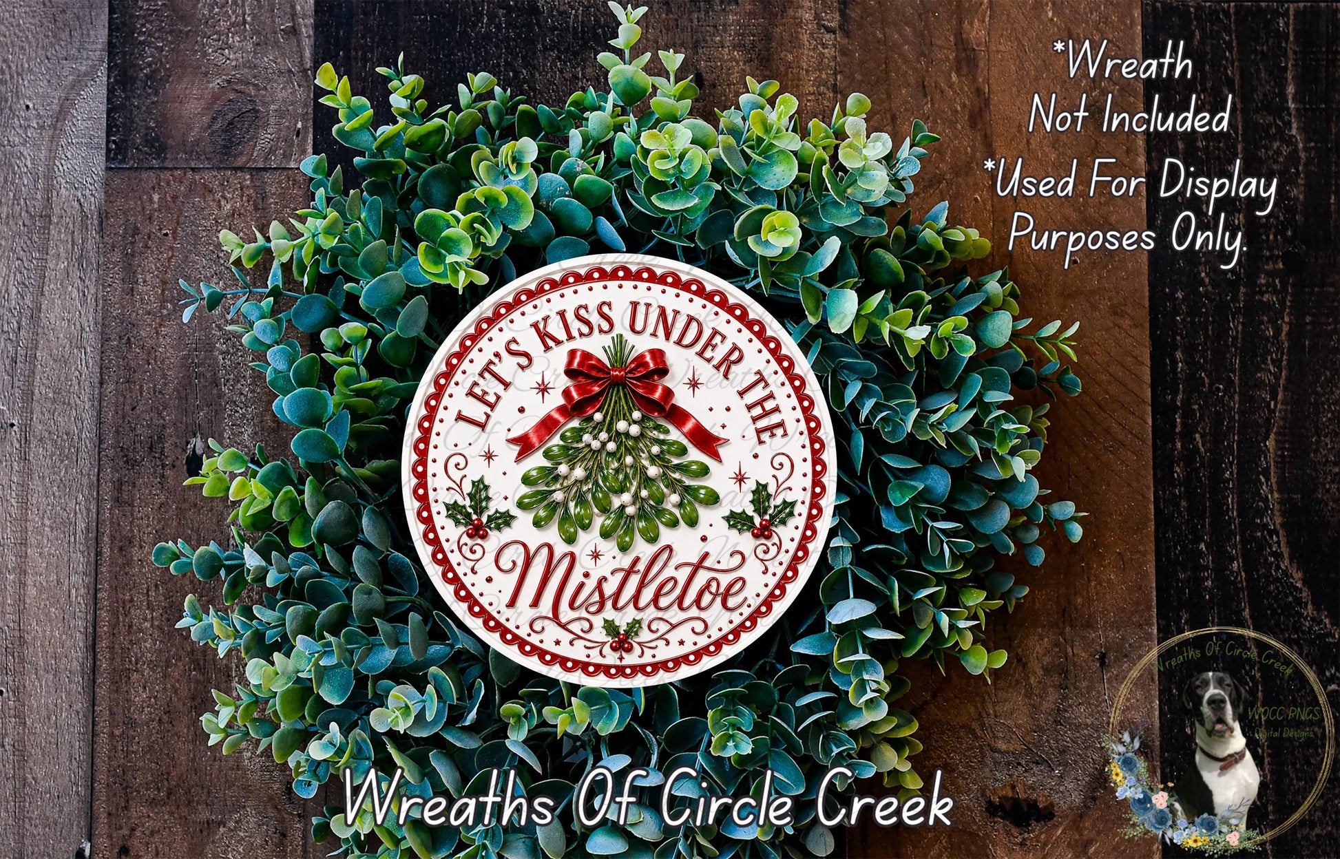 A wreath made of green leaves and a red ribbon, with a circular sign that says "Let's Kiss Under The Mistletoe" in the center.