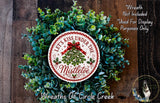 A wreath made of green leaves and a red ribbon, with a circular sign that says "Let's Kiss Under The Mistletoe" in the center.