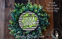 a wreath made of green and white leaves, with a central circular sign that says "SOMEBODY'S LUCKY CHARM". The wreath is displayed on a wooden surface, and there is a small dog in the bottom right corner of the image.