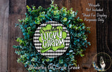 a wreath made of green and white leaves, with a central circular sign that says "SOMEBODY'S LUCKY CHARM". The wreath is displayed on a wooden surface, and there is a small dog in the bottom right corner of the image.