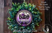 a wreath made of green leaves and a purple circular sign with the text "I put the BOO in BOUJE" on it. The sign is surrounded by a lush, wreath-like arrangement of leaves. The background is a wooden wall with a dog visible in the corner.