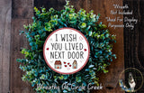 a wreath made of green leaves and a red and white sign with the text "I WISH YOU LIVED NEXT DOOR" displayed on it.