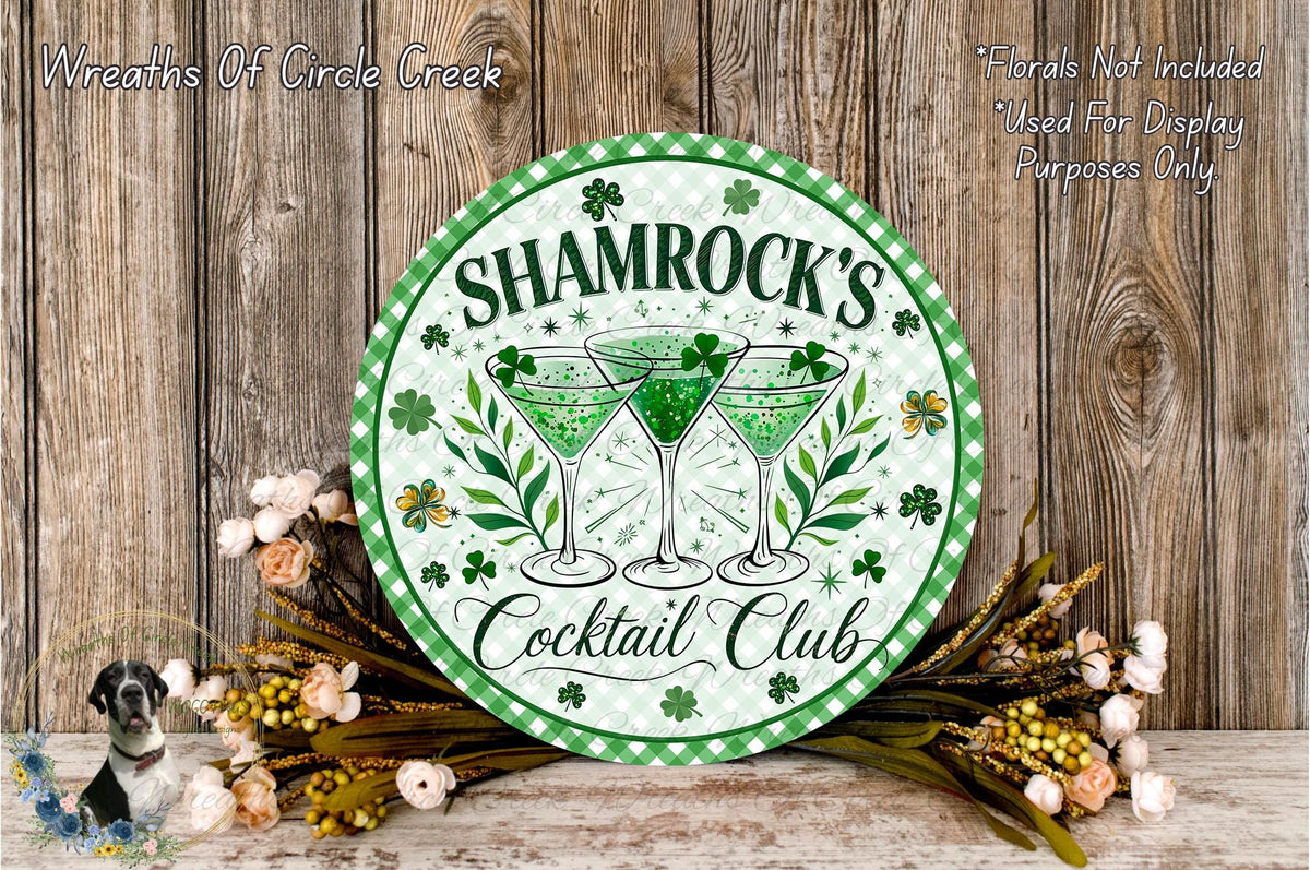 a circular sign with the text " Shamrock's Cocktail Club" and three martini glasses with shamrocks, surrounded by flowers and greenery.
