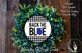 a wreath made of green leaves and a blue shield with the text "BACK THE BLUE" displayed on it. The wreath is placed on a wooden surface, and there is a small dog in the bottom right corner of the image.