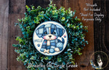 A wreath made of green plants and featuring a cartoon dog wearing a blue outfit with the text "#Hello" on it.