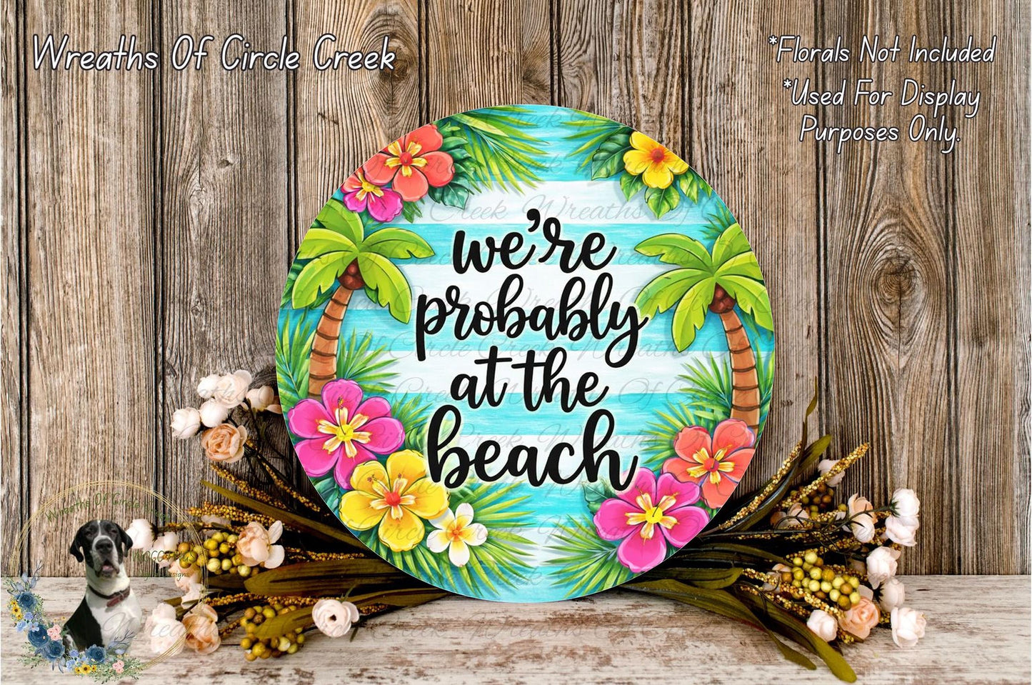 A round wooden sign with a tropical beach theme, featuring palm trees, flowers, and the text "we're probably at the beach". The sign is surrounded by a wreath of flowers and placed on a wooden surface.