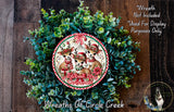 A Christmas wreath made of green foliage and featuring three deer wearing Santa hats. The deer are wearing red and white scarves and have antlers. The wreath is displayed on a wooden surface.