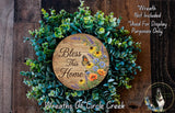 A wreath made of various green plants and flowers, with a wooden sign that says "Bless This Home" in the center.