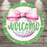A circular welcome sign with a pink bow, surrounded by a green border with golf balls and clubs. The sign displays the word "welcome" in a cursive font.