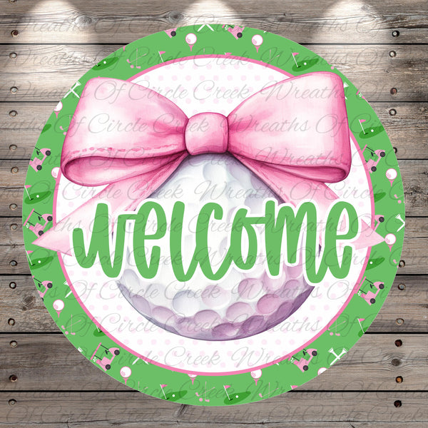 A circular welcome sign with a pink bow, surrounded by a green border with golf balls and clubs. The sign displays the word "welcome" in a cursive font.