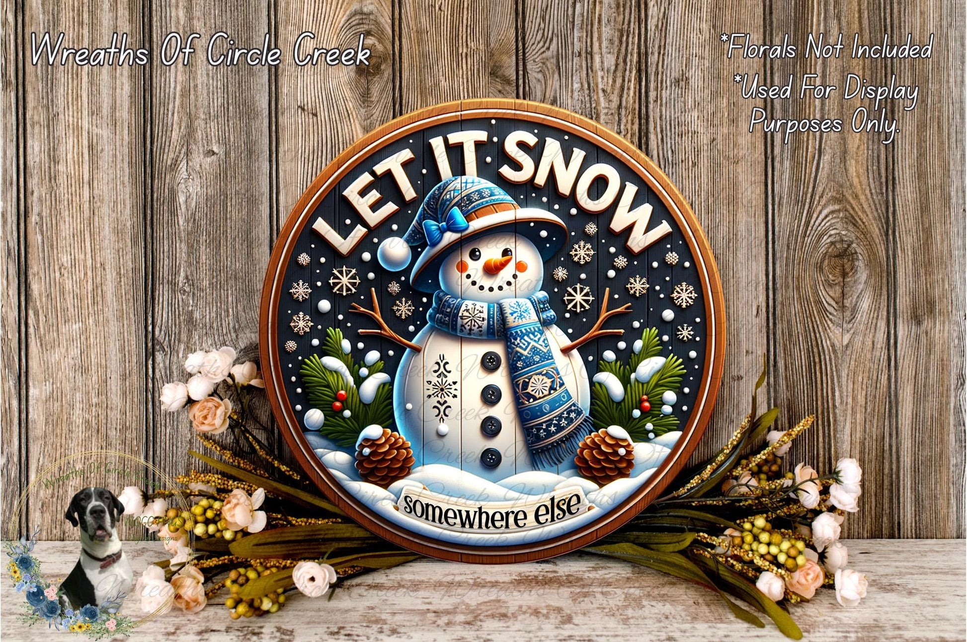 A decorative snowman sign with the text "LET IT SNOW somewhere else" surrounded by pine cones, pine branches, and flowers, placed on a wooden surface.