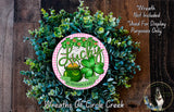 A wreath made of green leaves and a clover, with a pink and white circular sign featuring the word "Lucky" and an illustration of a pot of gold.