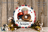 a round sign with a baseball theme, surrounded by flowers and a baseball glove. The sign displays the text "Tis the Season" and includes a red popcorn bucket and a baseball. The sign is set against a wooden background, and there is a small bird figurine in the bottom left corner.