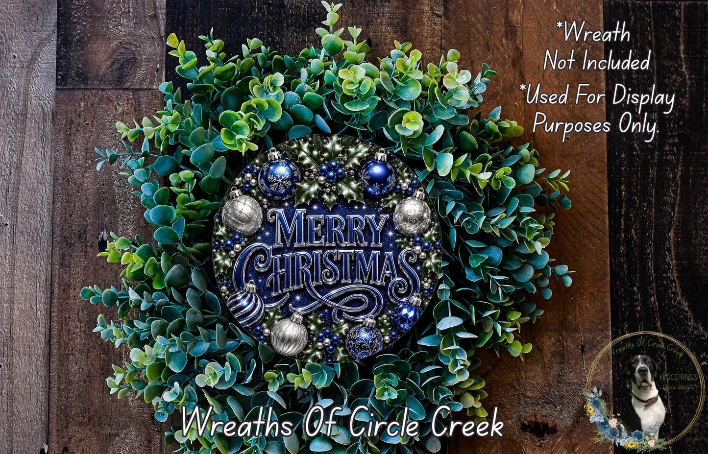 A wreath made of green and blue foliage, with a blue and silver ornament in the center, and the text "Merry Christmas" written on it. The wreath is displayed on a wooden surface, and there is a small dog in the bottom right corner of the image.
