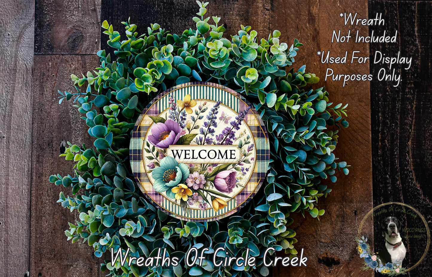 a wreath made of various green plants and flowers, with a circular sign in the center that says "WELCOME". The wreath is displayed on a wooden surface, and there is a small dog in the bottom right corner of the image.