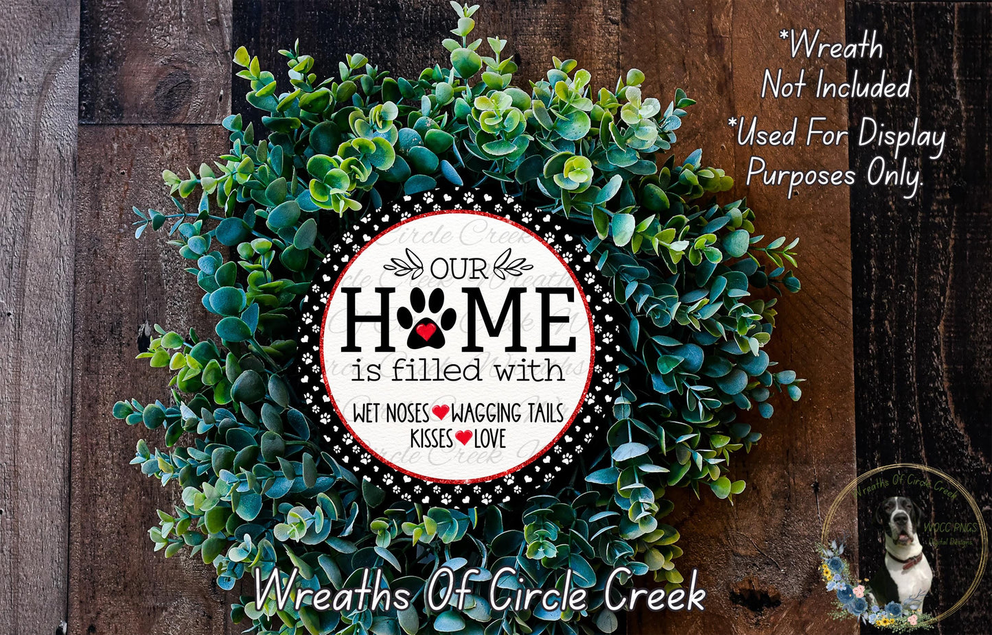 A wreath made of green leaves and a circular sign with the text "OUR HOME is filled with WET Noses WAGING Tails KISSES LOVE" is displayed on a wooden surface.