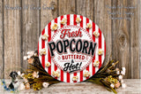 a circular sign with the text "Fresh Popcorn Buttered & Hot!" surrounded by popcorn and flowers, placed on a wooden surface.