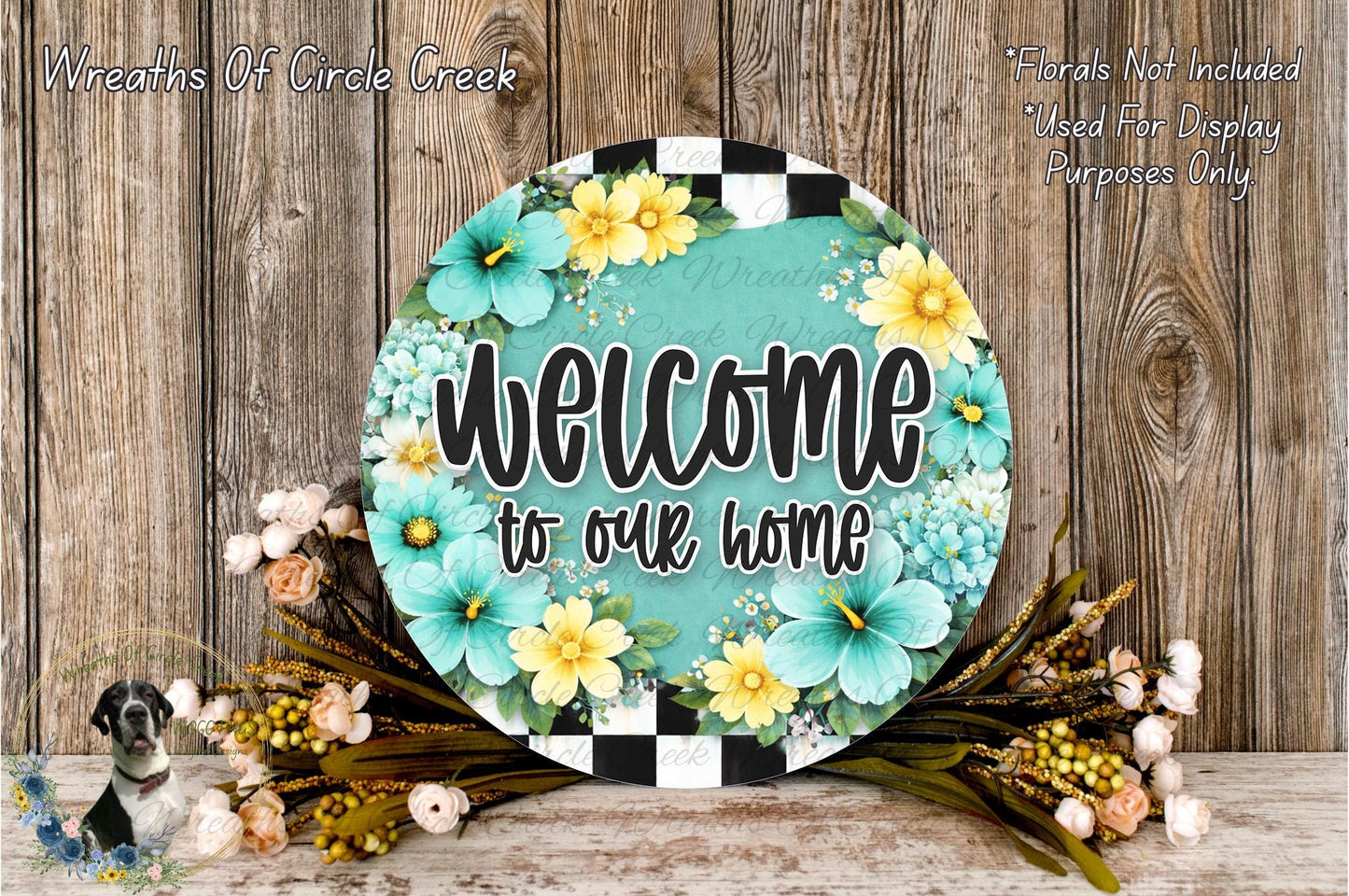 a round sign with the text "Welcome to our home" surrounded by a floral arrangement. The sign is placed on a wooden surface, and there are flowers and greenery surrounding it.