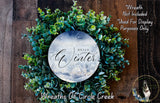 A wreath made of green leaves and flowers, with a circular sign displaying the text "Hello Winter" in the center.