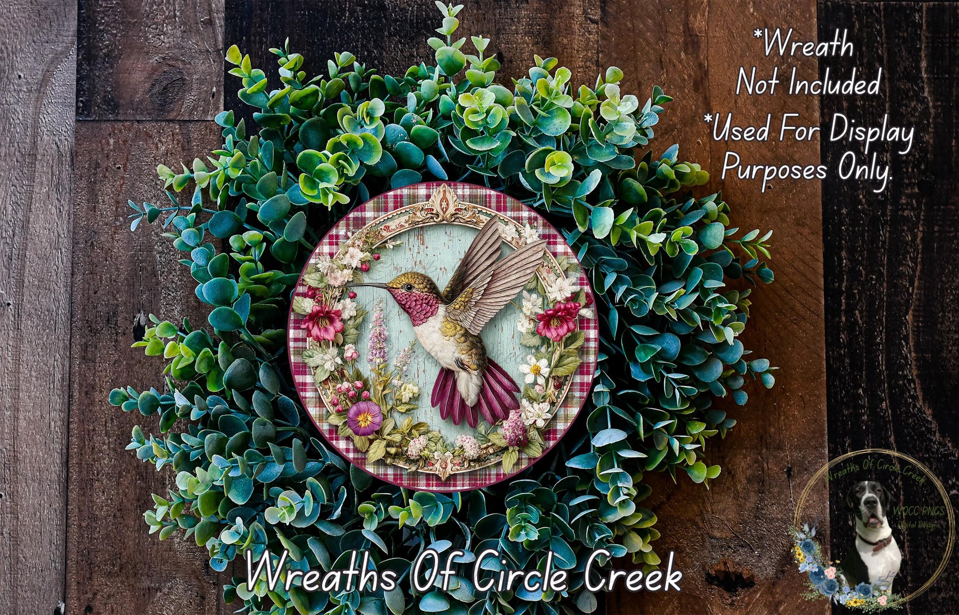 a decorative wreath with a hummingbird on it, surrounded by various green plants and flowers. The wreath is placed on a wooden surface, and there is a small dog visible in the bottom right corner of the image.