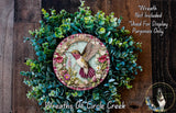 a decorative wreath with a hummingbird on it, surrounded by various green plants and flowers. The wreath is placed on a wooden surface, and there is a small dog visible in the bottom right corner of the image.