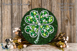 A decorative round wreath made of green glitter and rhinestones, with a four-leaf clover design, surrounded by flowers and placed on a wooden surface.