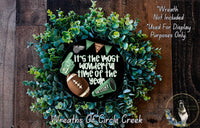 A wreath made of green foliage, with a football and cheerleading gear displayed on it. The wreath is surrounded by text that reads "It's the most wonderful time of the year" and "It's the most wonderful time of the year".