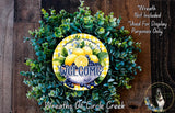 a wreath made of green leaves and yellow flowers, with a yellow and blue sign that says "Welcome" in the center. The wreath is displayed on a wooden surface, and there is a small dog in the bottom right corner.