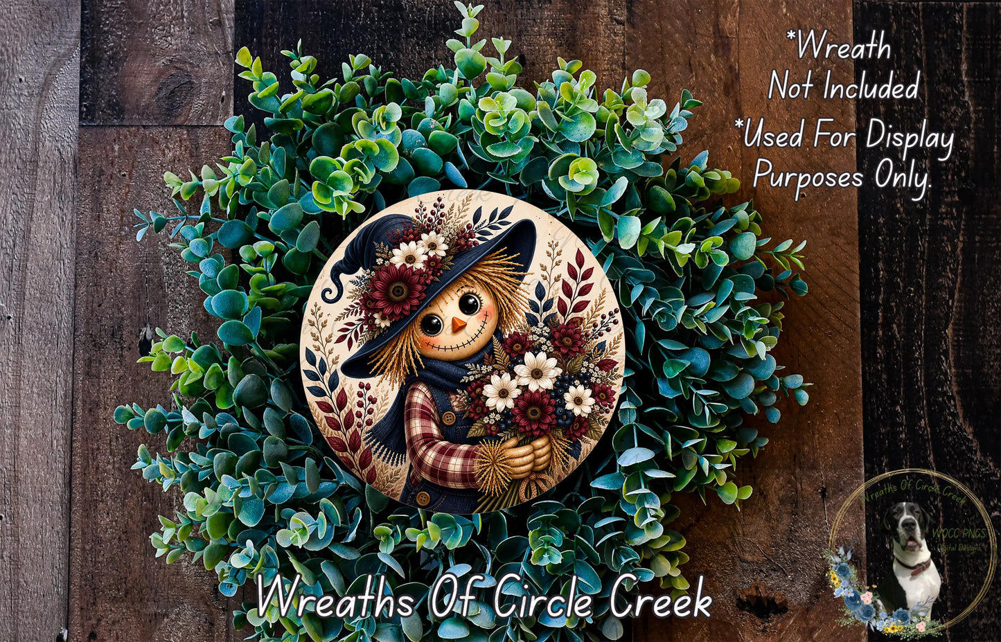 a wreath with a scarecrow figure holding flowers, surrounded by a lush green wreath. The wreath is displayed on a wooden surface, and there is a small dog in the bottom right corner of the image.