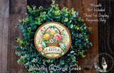 a wreath made of various green plants and flowers, with a decorative plate in the center that says "Happy Easter". The wreath is displayed on a wooden surface, and there is a small dog in the bottom right corner of the image.