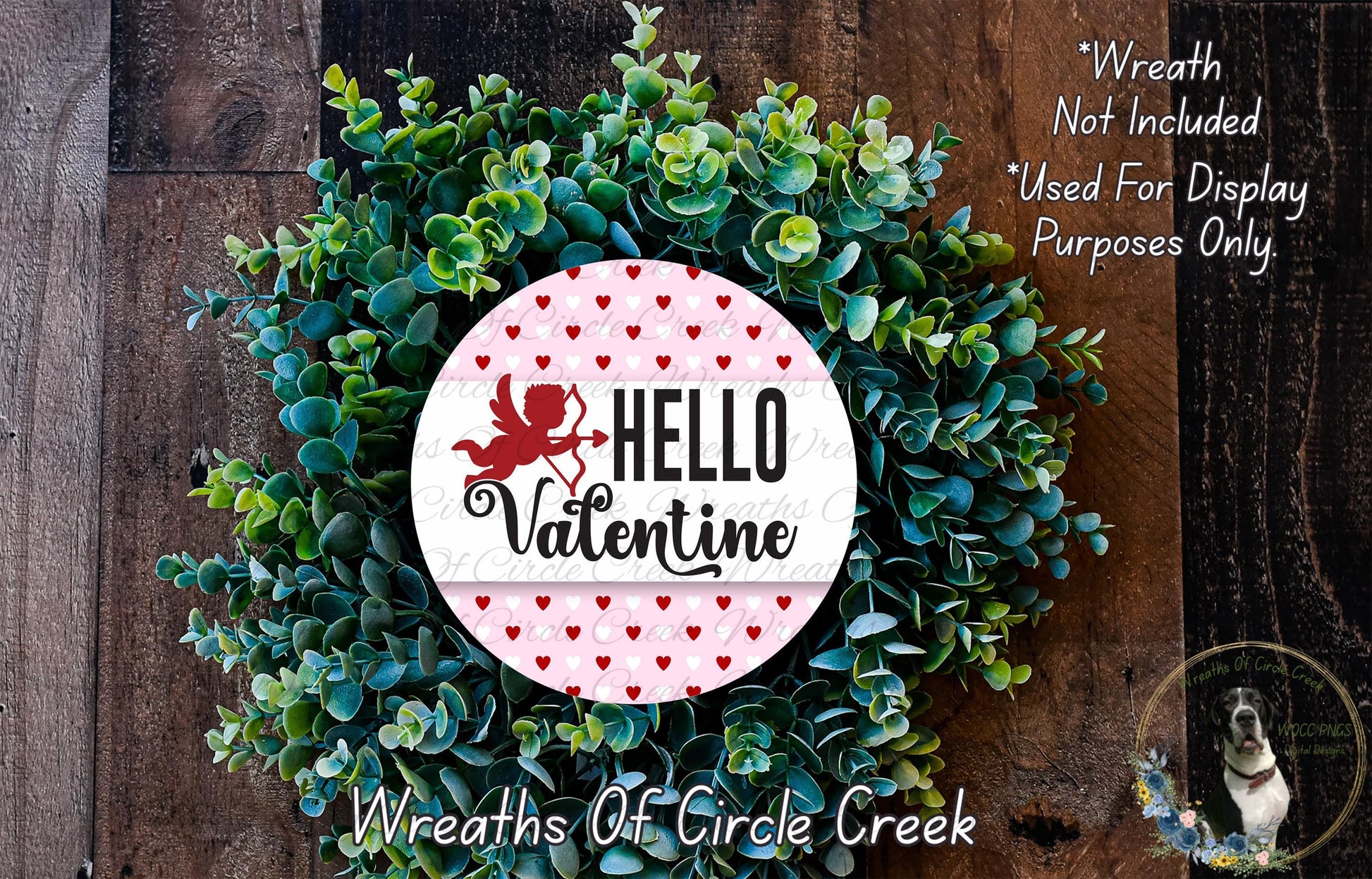 a wreath made of green leaves and a red heart-shaped ornament with the text "Hello Valentine" on it. The wreath is displayed on a wooden surface, and there is a small dog in the bottom right corner of the image.