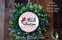 a wreath made of green leaves and a red heart-shaped ornament with the text "Hello Valentine" on it. The wreath is displayed on a wooden surface, and there is a small dog in the bottom right corner of the image.