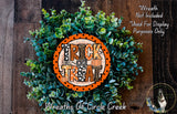 a wreath made of green foliage and an orange circular sign with the text "TRICK TREAT" on it, placed on a wooden surface.