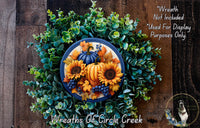 A wreath made of various fall-themed elements, including sunflowers, pumpkins, and berries, is displayed on a wooden surface.