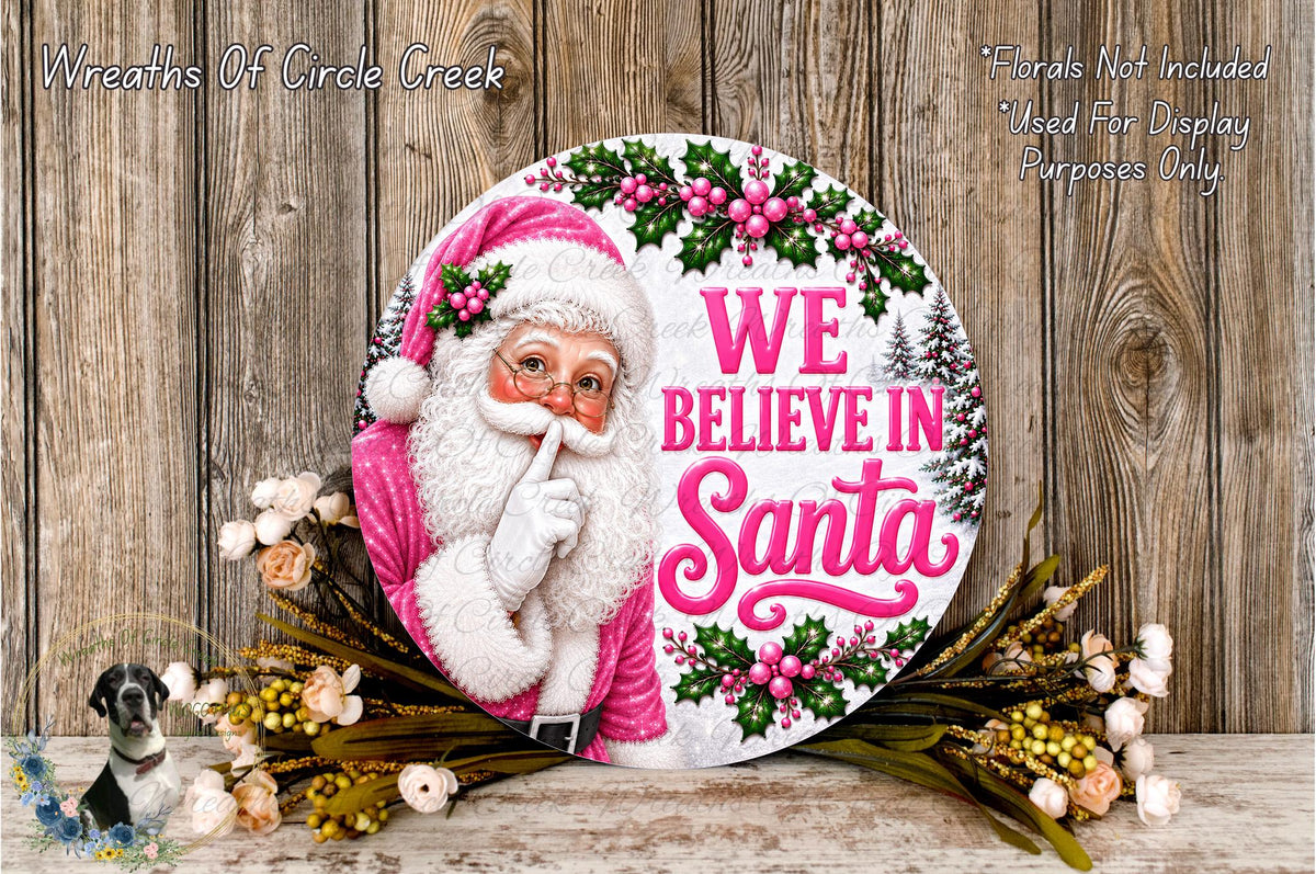 A round sign featuring a Santa Claus figure with the text "WE BELIEVE IN Santa" and surrounded by flowers and greenery.