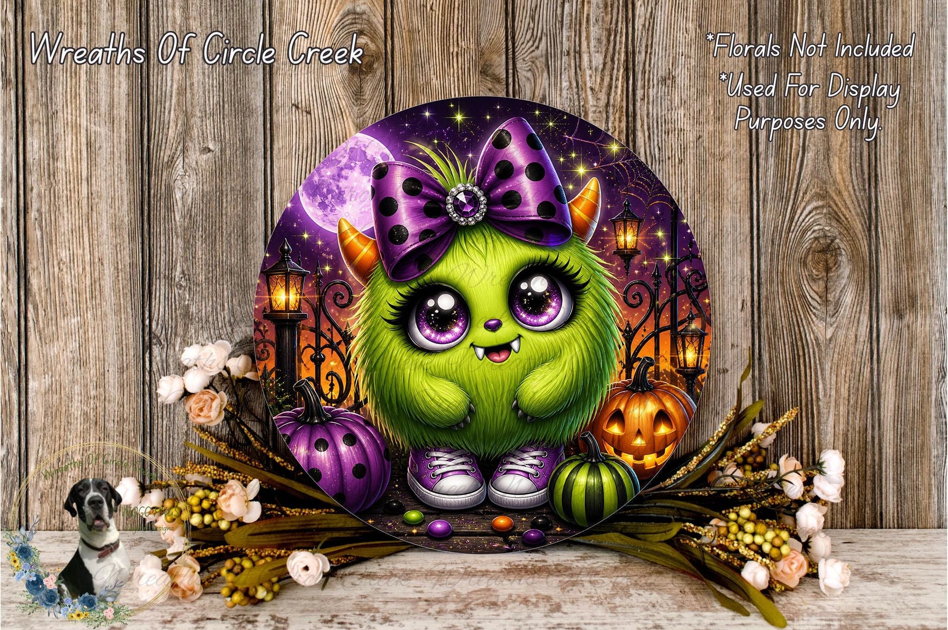 A whimsical, cartoonish green monster with large eyes and a purple bow sits amidst Halloween-themed decorations, including pumpkins, lanterns, and flowers, against a wooden background.