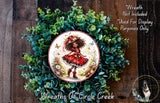 A wreath made of green leaves and flowers, with a circular plate featuring a picture of a young girl wearing a red dress and a flower crown.