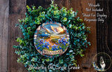 a wreath with a welcome sign, surrounded by greenery and flowers, with a scenic mountain landscape in the background.