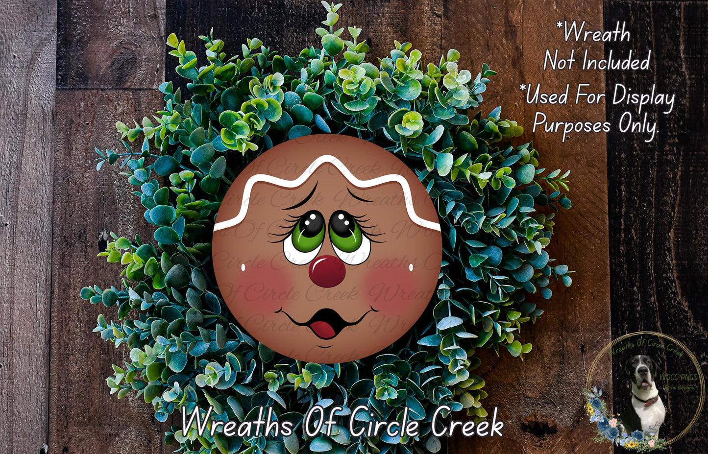 A wreath made of green plants and a gingerbread man-shaped cookie with a red nose and green eyes, placed on a wooden surface.