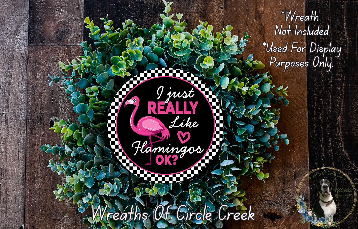 a wreath made of green foliage and a pink flamingo, with a humorous text overlay that reads "I just really like flamingos, OK?". The wreath is displayed on a wooden surface, and there is a small dog visible in the bottom right corner of the image.