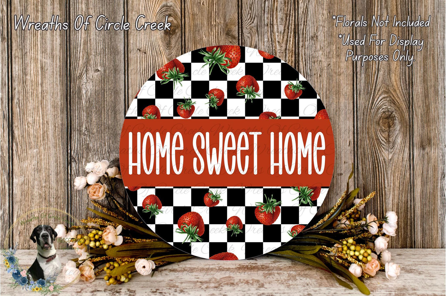 a round sign with the text "HOME SWEET HOME" in bold red letters, surrounded by a checkered pattern of red and white tomatoes. The sign is placed on a wooden surface, with a floral arrangement of flowers and dried leaves in the foreground.
