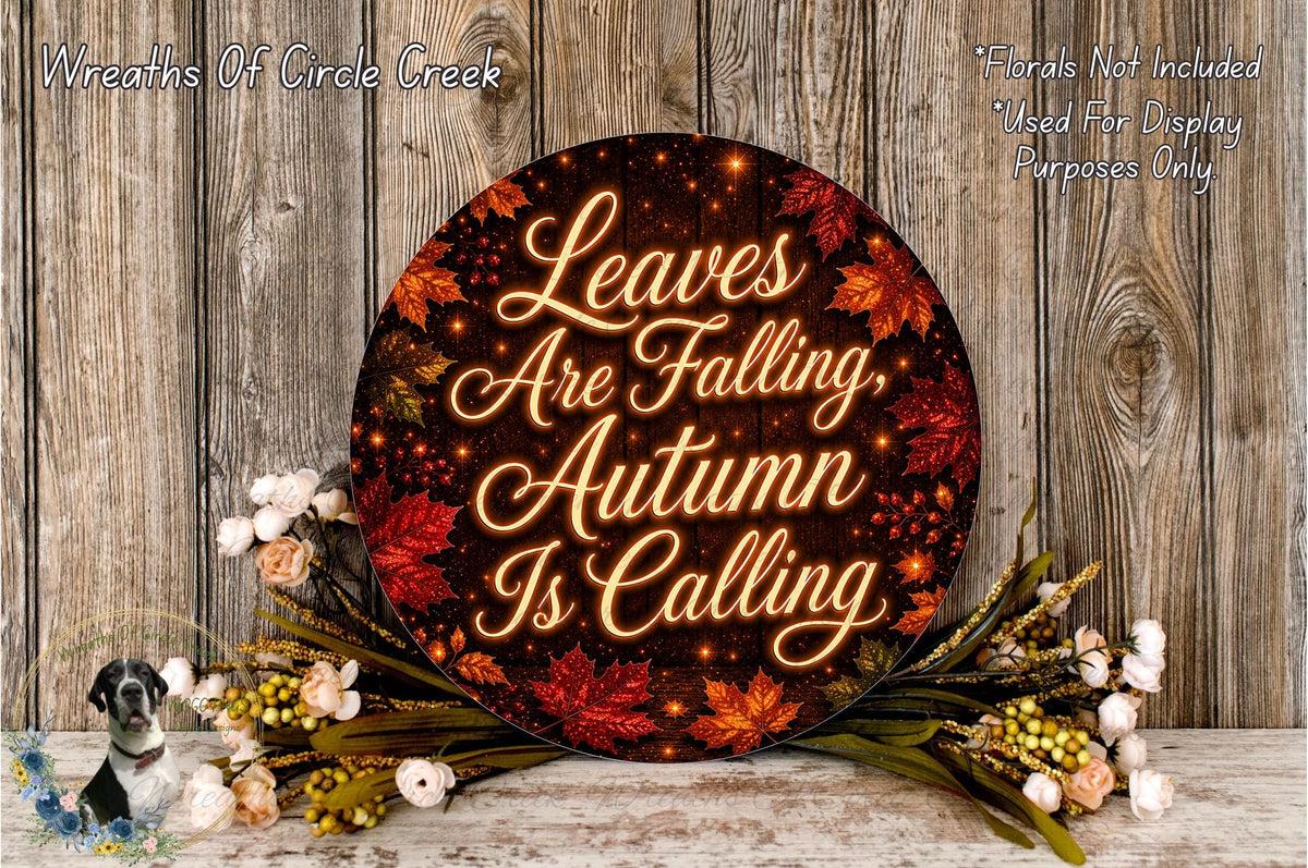 a circular sign with the text "Leaves Are Falling, Autumn Is Calling" surrounded by autumn leaves and flowers, placed on a wooden surface.