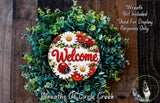 a wreath with a welcome sign in the center, surrounded by various green leaves and flowers. The wreath is displayed on a wooden surface, and there is a small dog in the bottom right corner of the image.