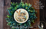 A wreath featuring two rabbits wearing hats and holding flowers, surrounded by a lush green plant.