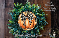 a wreath made of various green plants and a large orange pumpkin with a black and white checkered bow, hanging on a wooden wall.