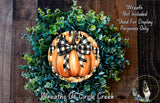 a wreath made of various green plants and a large orange pumpkin with a black and white checkered bow, hanging on a wooden wall.