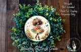 a wreath with a central image of a fairy wearing a colorful dress and hat, surrounded by a lush green wreath. The wreath is displayed on a wooden surface, and there is a small dog in the bottom right corner of the image.