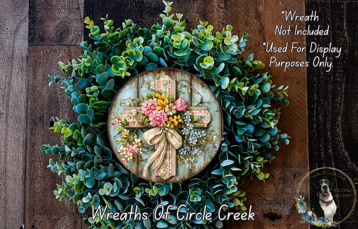 a wreath made of various green plants and flowers, including a cross at the center. The wreath is displayed on a wooden surface, and there is a small dog in the bottom right corner of the image.