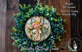 a wreath made of various green plants and flowers, including a cross at the center. The wreath is displayed on a wooden surface, and there is a small dog in the bottom right corner of the image.