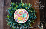 a wreath made of green leaves and flowers, with a colorful sign that reads "Autism Mom" in the center. The wreath is displayed on a wooden surface, and there is a small dog in the bottom right corner of the image.